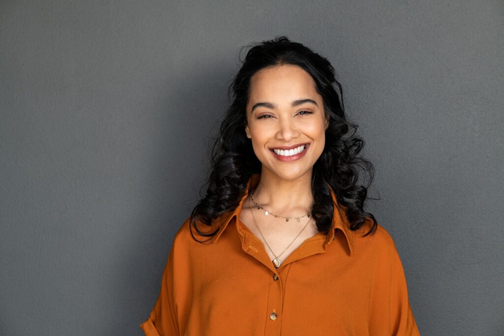 Confident Woman Smiling - Dentist in San Fernando A cheerful woman with curly hair in a burnt orange blouse smiling confidently against a gray background - Dentist in San Fernando