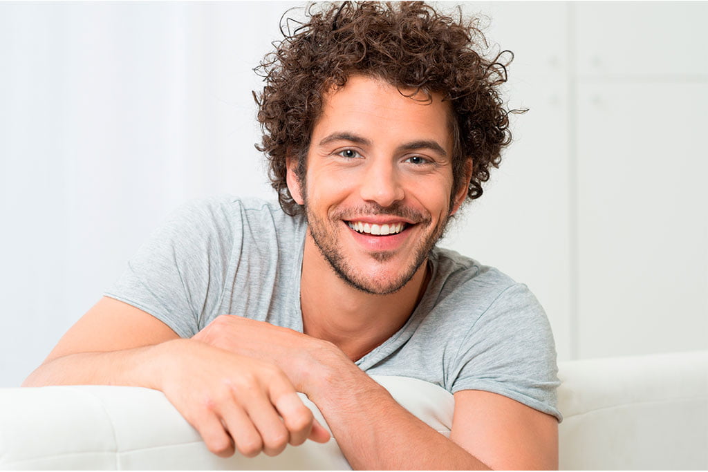 Young Man with Healthy Smile - Dentist in San Fernando A cheerful young man with curly hair smiling while leaning on a couch - Dentist in San Fernando