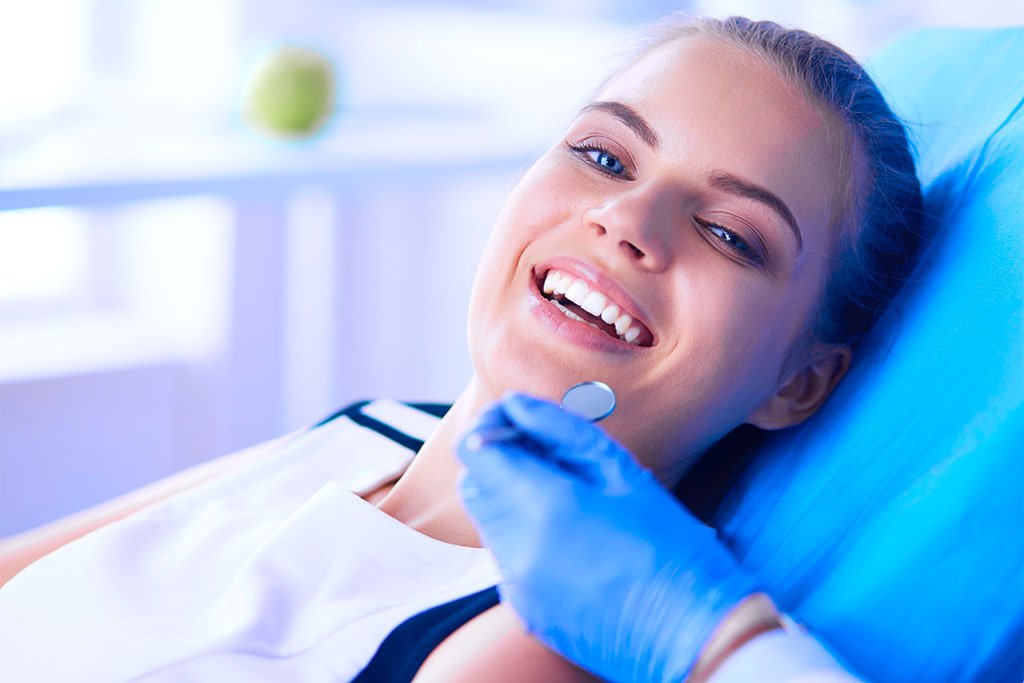 Relaxed Woman at Dental Appointment - Dentist in San Fernando A young woman reclining in a dental chair smiling while being examined with a dental mirror - Dentist in San Fernando