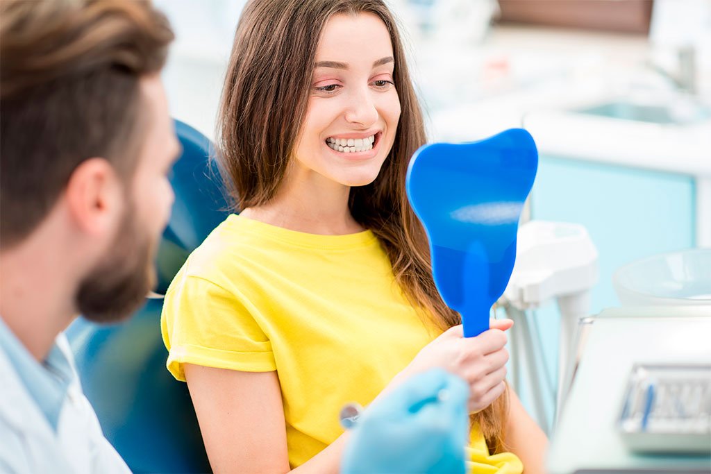 Patient Checking Smile in Mirror - Dentist in San Fernando A female dental patient smiling into a blue tooth-shaped mirror during her appointment - Dentist in San Fernando