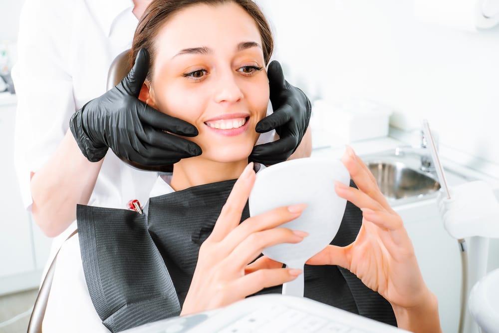 Woman Checking Her Smile After Dental Treatment – Dental Implants in San Fernando, CA A woman sits in the dental chair holding a mirror while a dental professional adjusts her smile, highlighting her satisfaction with the results – Dental Implants in San Fernando, CA.
