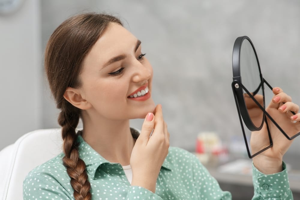 Woman Admiring Her New Smile in Mirror – Veneers San Fernando CA A smiling woman checks her reflection in a handheld mirror, appreciating the natural-looking results of her newly improved smile – Veneers San Fernando CA