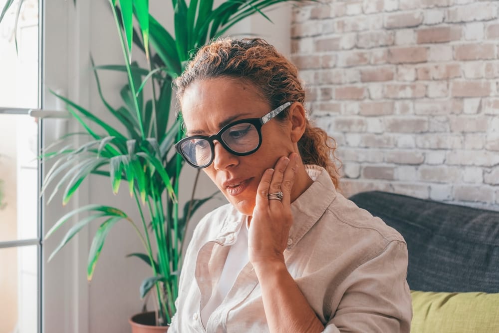 A woman sits at home holding her cheek with visible discomfort, indicating possible tooth pain or a dental emergency requiring urgent care – Emergency Dentist in San Fernando, CA.