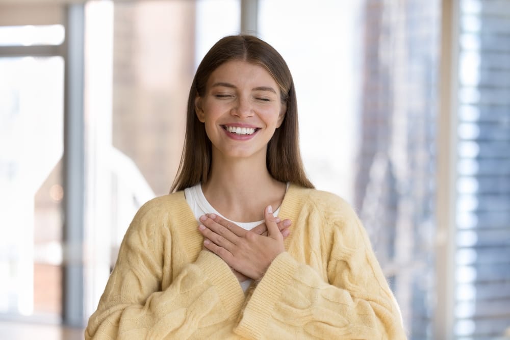 A serene young woman embraces a moment of gratitude with her hands placed on her chest, eyes closed, and a bright smile that reflects happiness and wellness – Dentist in Pacoima, CA.