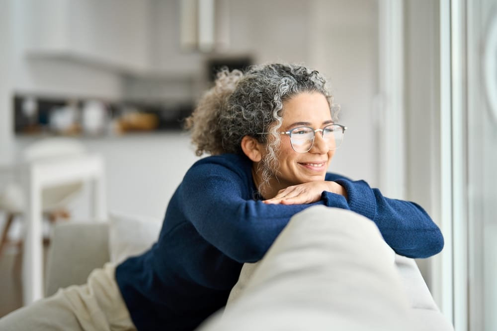A serene older woman with curly gray hair and glasses leans comfortably on her couch, smiling softly while enjoying a peaceful moment at home – Dentist in Sylmar, CA.