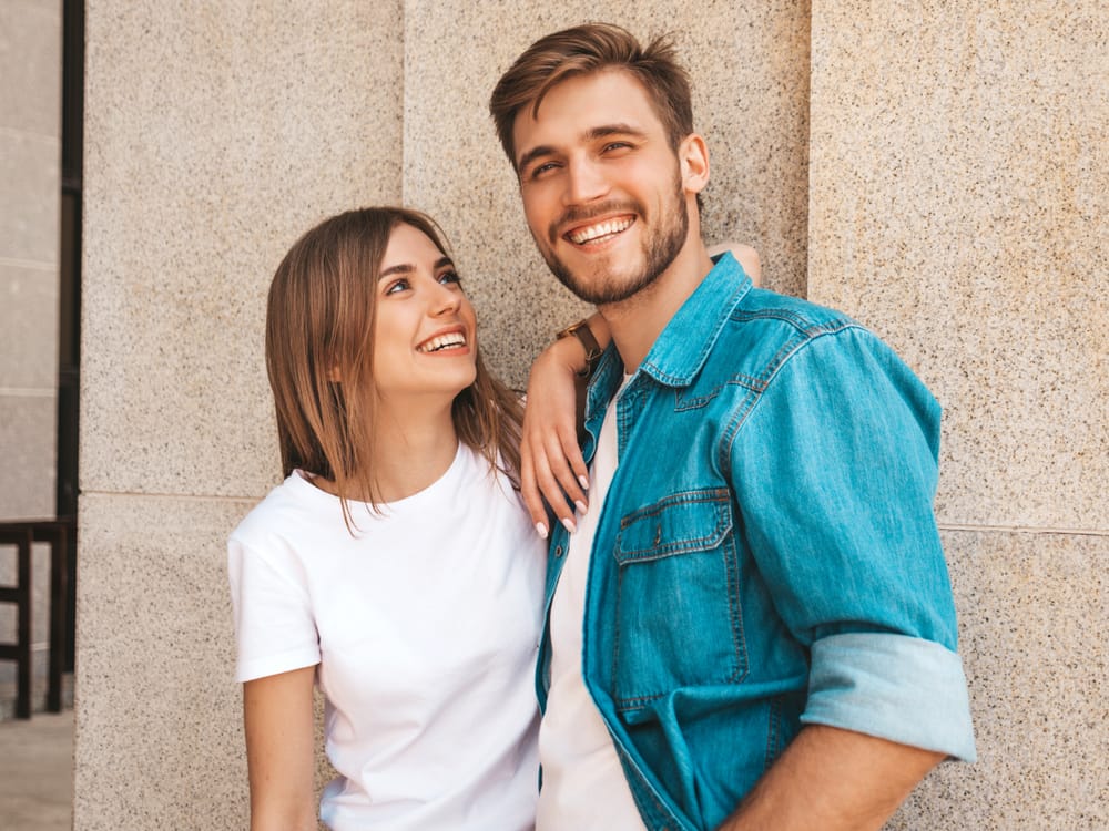 A cheerful couple stands close together, smiling brightly while leaning against a light stone wall, capturing a moment of connection and confidence – Dentist in Pacoima, CA.